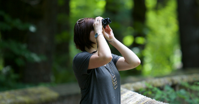 Woman looking into a pair of binoculars in the woods. Photo: Kayla Farmer, Unsplash.