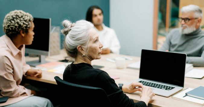 Four people sitting at a table, and a women in a black shirt sitting in front of a laptop. Photo: Cottonbro, from Pexels.