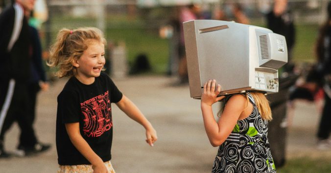 Two little girls facing each other. One of them is smiling and dancing, the other one has an old TV screen on her head. Photo: Ryunosuke Kikuno, from Unsplash.