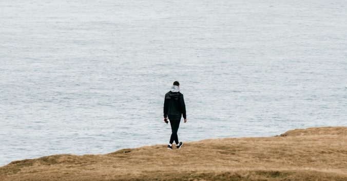 Man in dark clothes walking alone on a hill beside the ocean. Photo: Polina Kuzovkova, from Unsplash.